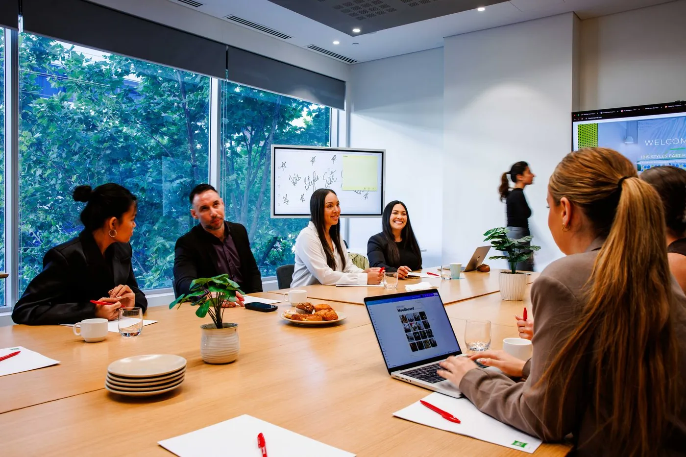 A team meeting around the conference table, laptops open, with trees outside the full-height windows