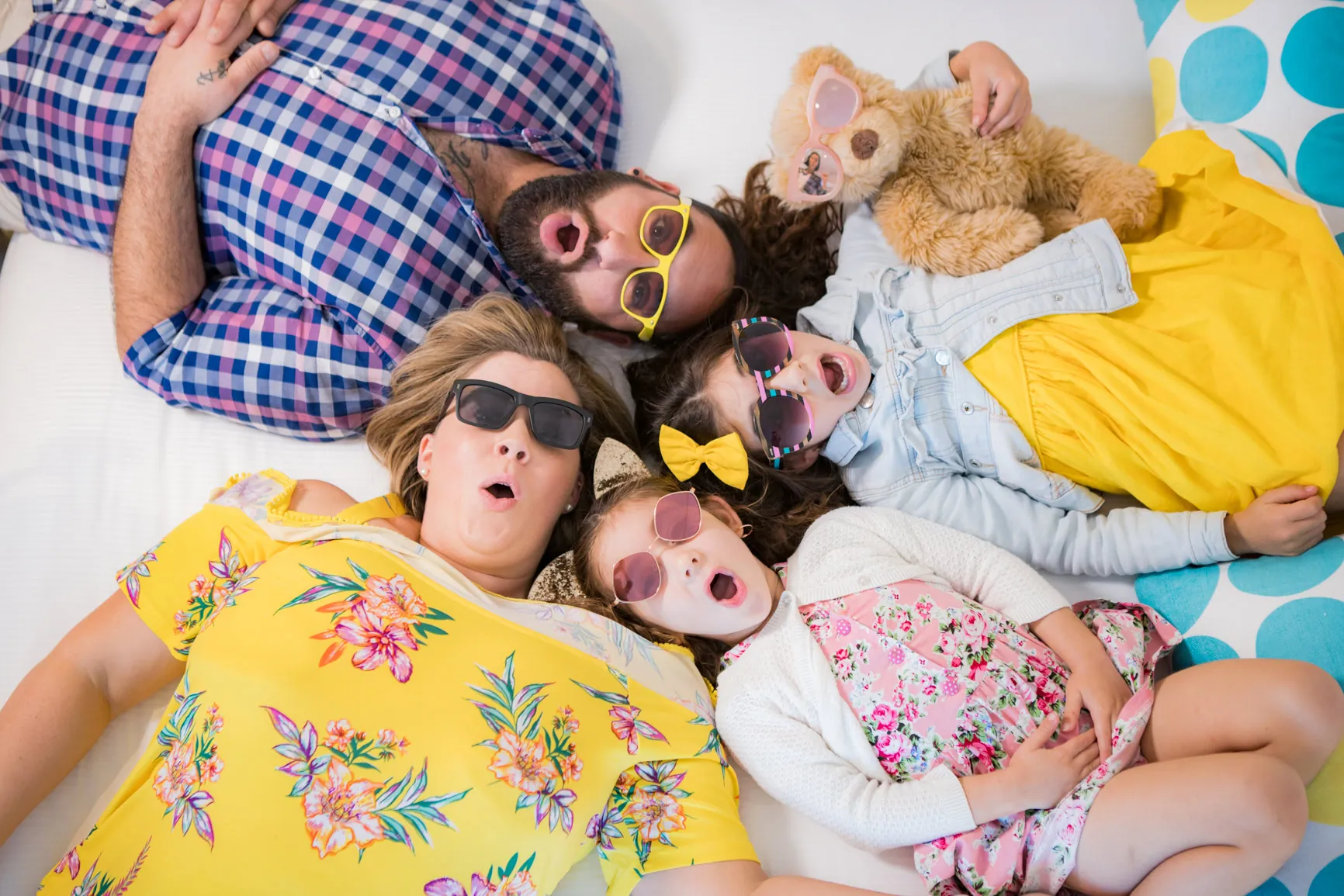 A family lying on a bed in colourful clothes and playful sunglasses, smiling, the ibis Styles creative, fun-loving spirit