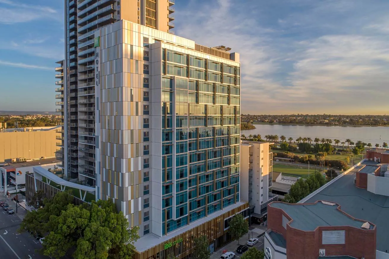 The ibis Styles East Perth hotel tower at dusk, its patterned facade lit by the low sun, with the Swan River and the foreshore behind