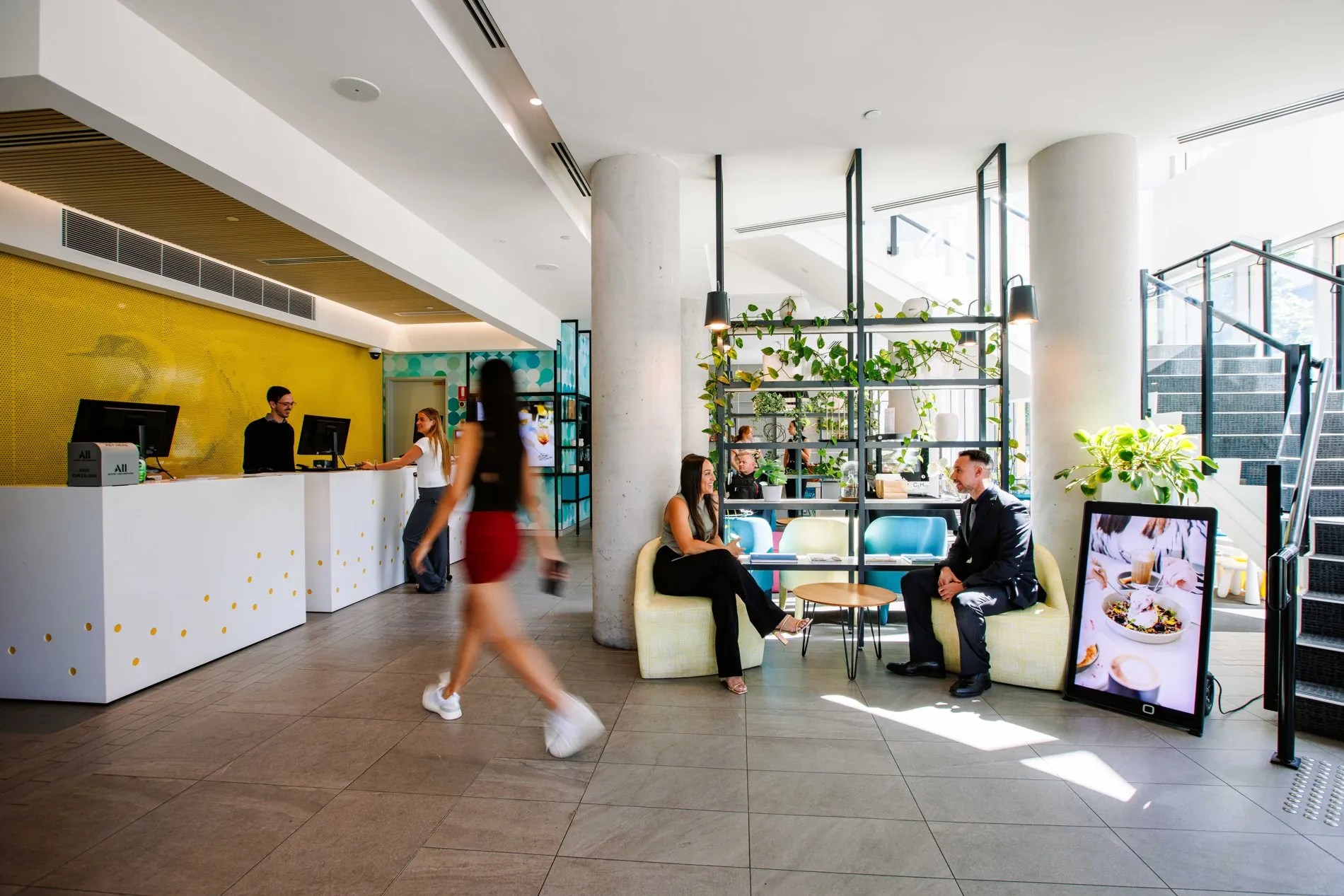 The ibis Styles East Perth lobby: guests checking in at the white desk against the bright yellow wall, while others sit and talk in the green and blue lounge chairs among hanging plants