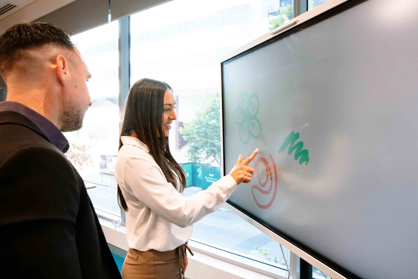 Two colleagues drawing on the interactive screen in a meeting room, the city outside the window