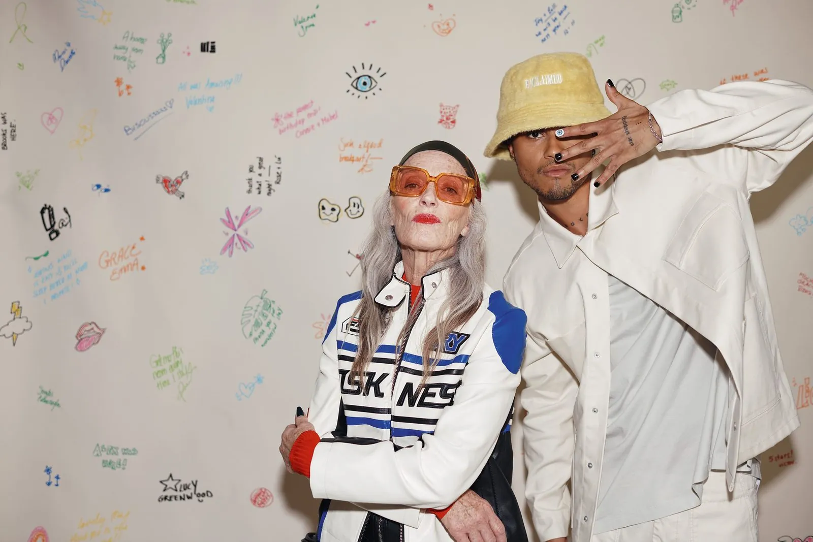 An older guest in orange sunglasses and a bold striped jacket standing arm in arm with a younger guest against the ibis Styles doodle wall