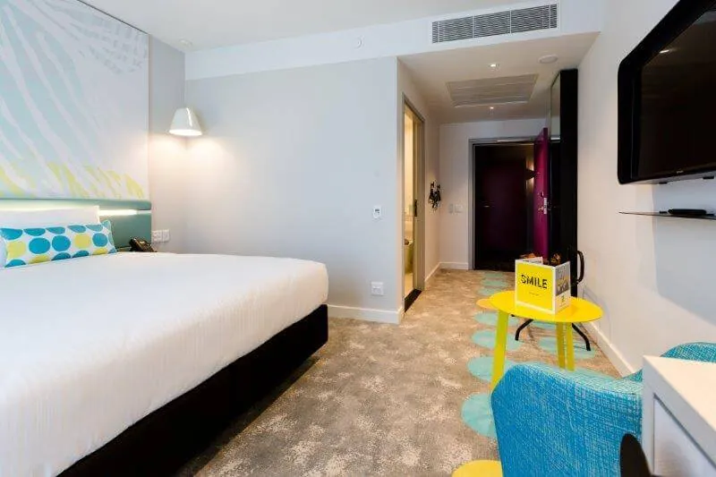 An accessible King room: a king bed under the mural wall with a wide, clear passage running past the foot of the bed to the door, a blue armchair and a yellow side table on the right