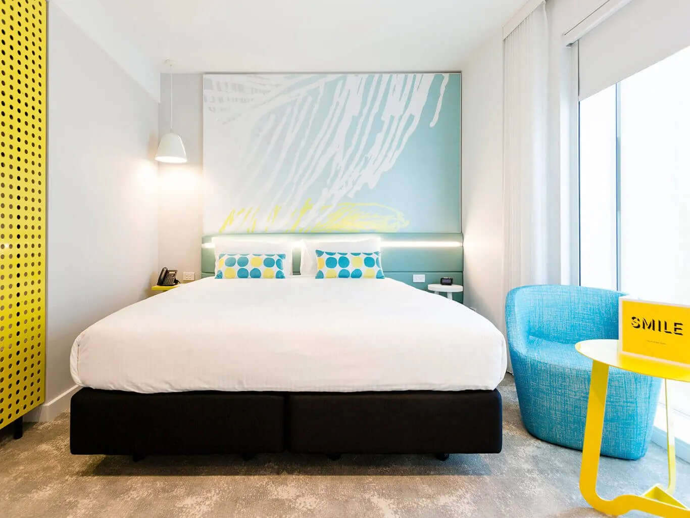 A Deluxe King room: a king bed under the turquoise mural wall, a yellow perforated screen on the left and a blue armchair with a yellow side table by the window