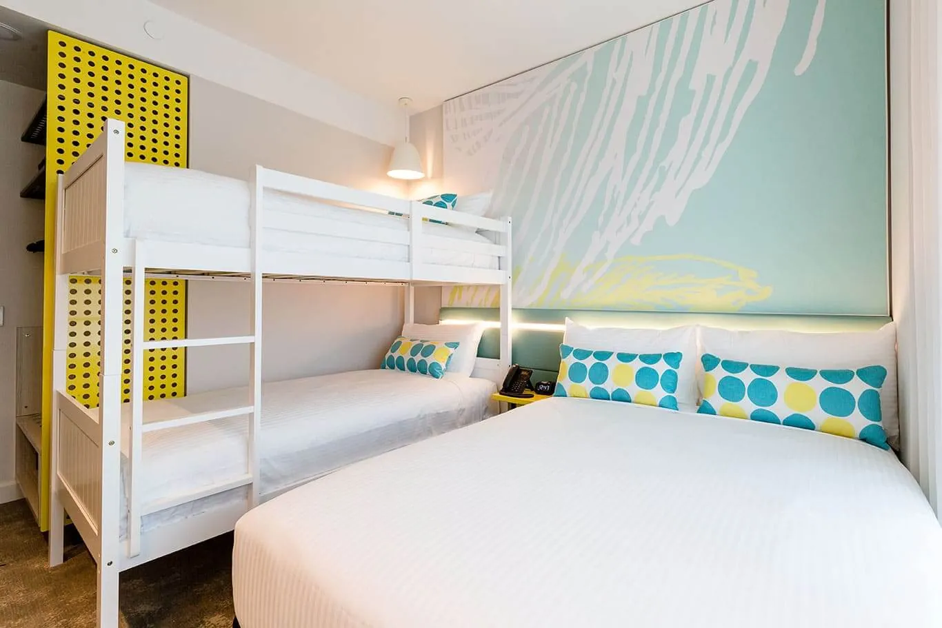 A Family room: a double bed with spotted cushions beside a white bunk bed, the turquoise mural wall behind and a yellow perforated screen by the door