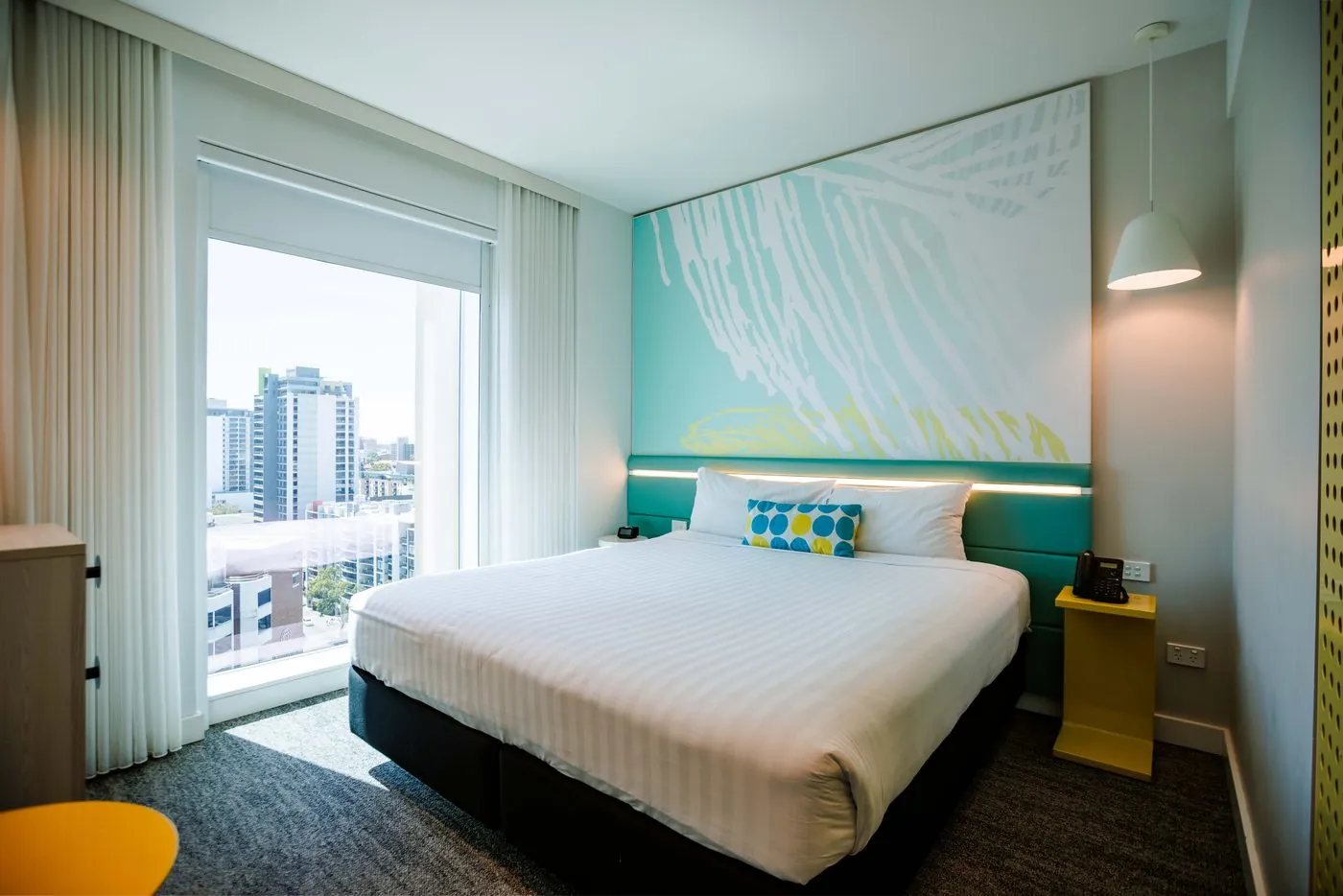 A Superior King room: a king bed with a spotted cushion under the turquoise mural wall, a yellow bedside table, and the East Perth skyline through the full-height window
