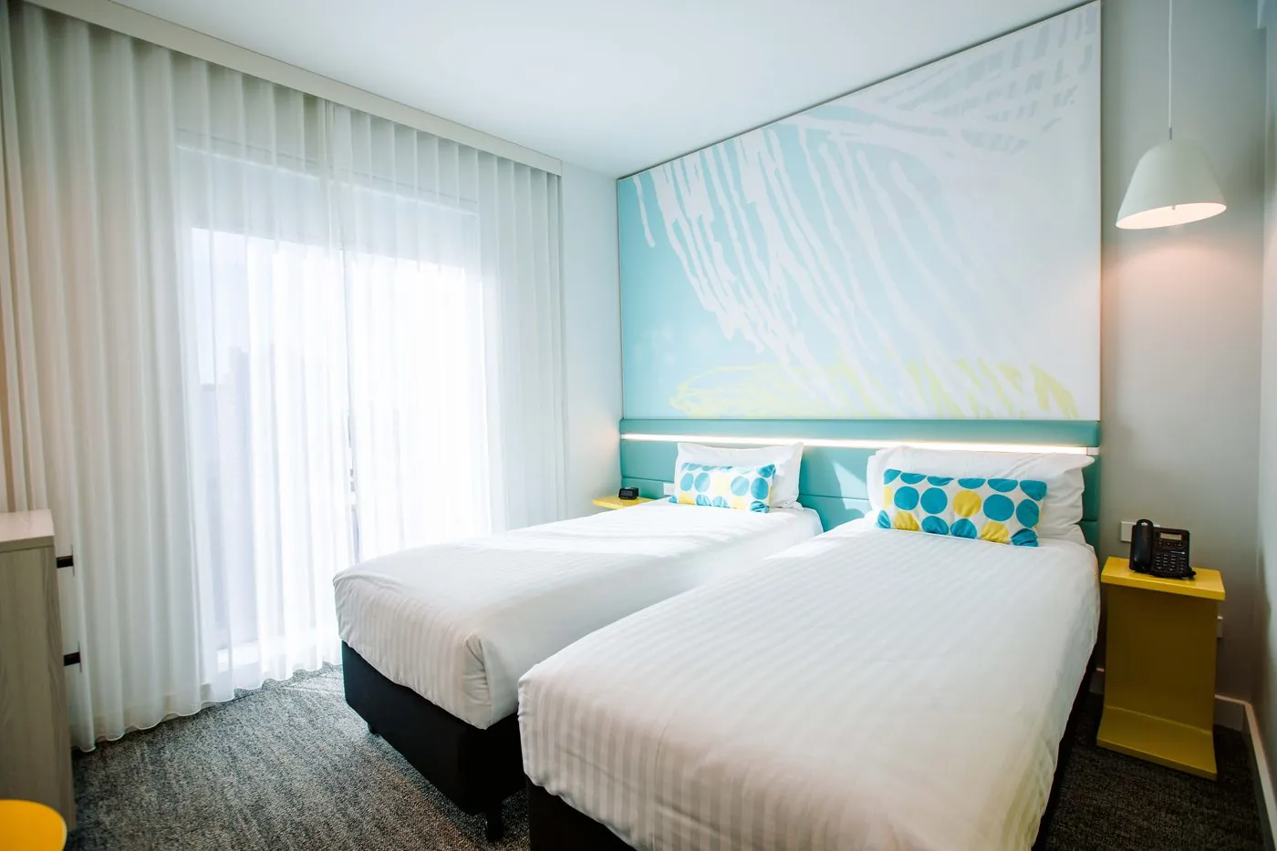 A Standard Twin room: two single beds with blue-and-yellow spotted cushions under the turquoise mural wall, a yellow bedside table and sheer curtains over the full-height window