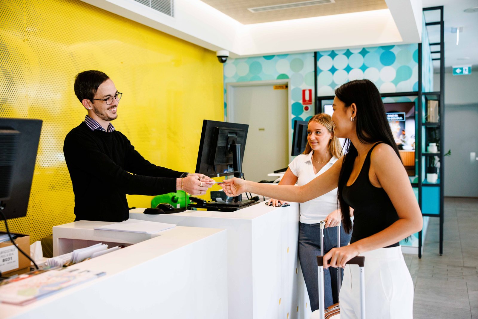 A receptionist handing a key card across the desk to two arriving guests, the yellow perforated wall behind him